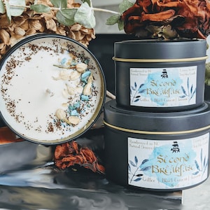 May include: Two black candle jars with gold lids. The larger jar has a white candle with brown and blue glitter and white flower petals. The smaller jar has a label that reads "Second Breakfast" with a watercolor design of blue and green leaves. The label also says "Coffee | Black Currant | Jasmine 4 oz."