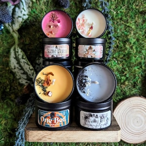 Peut inclure: Une collection de bougies artisanales dans des boîtes noires, chacune avec un parfum unique et des éléments décoratifs. Les bougies présentent diverses couleurs, dont le rose, le blanc, le jaune et le gris, avec des étiquettes portant les textes "Slow Burn", "Love Triangle", "One Bed" et "Morally Grey".