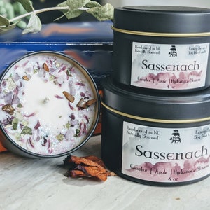 May include: Two black candle tins with gold trim, labeled "Sassenach" and featuring a watercolor design. The larger tin is 8 ounces and the smaller tin is 4 ounces. Both candles are hand-poured in North Carolina and naturally sourced. The scent is Camphor, Apple, and Hydrangea Blossom.