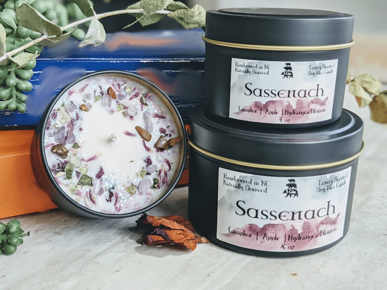 May include: Two black candle tins with gold trim, labeled "Sassenach" and featuring a watercolor design. The larger tin is 8 ounces and the smaller tin is 4 ounces. Both candles are hand-poured in North Carolina and naturally sourced. The scent is Camphor, Apple, and Hydrangea Blossom.