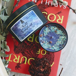May include: A black tin candle with a purple and blue galaxy design. The candle is labeled "Night Triumphant" with the scent notes "Rain | Citrus | Sea Salt". The candle is sitting on a red book with the title "A Court of Thorns and Roses" by Sarah J. Maas.