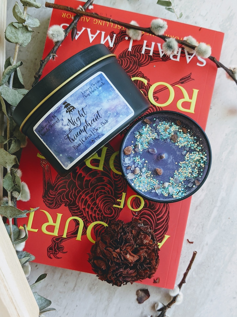 May include: A black tin candle with a purple and blue galaxy design. The candle is labeled "Night Triumphant" with the scent notes "Rain | Citrus | Sea Salt". The candle is sitting on a red book with the title "A Court of Thorns and Roses" by Sarah J. Maas.