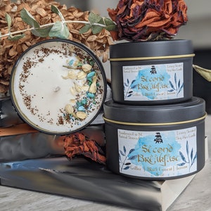 May include: Two black candle jars with gold lids. The smaller jar is on top of the larger jar. Both jars have a white label with black text that reads "Second Breakfast" and "Coffee | Black Current | Jasmine". The larger jar is 8 oz. The smaller jar is 4 oz.