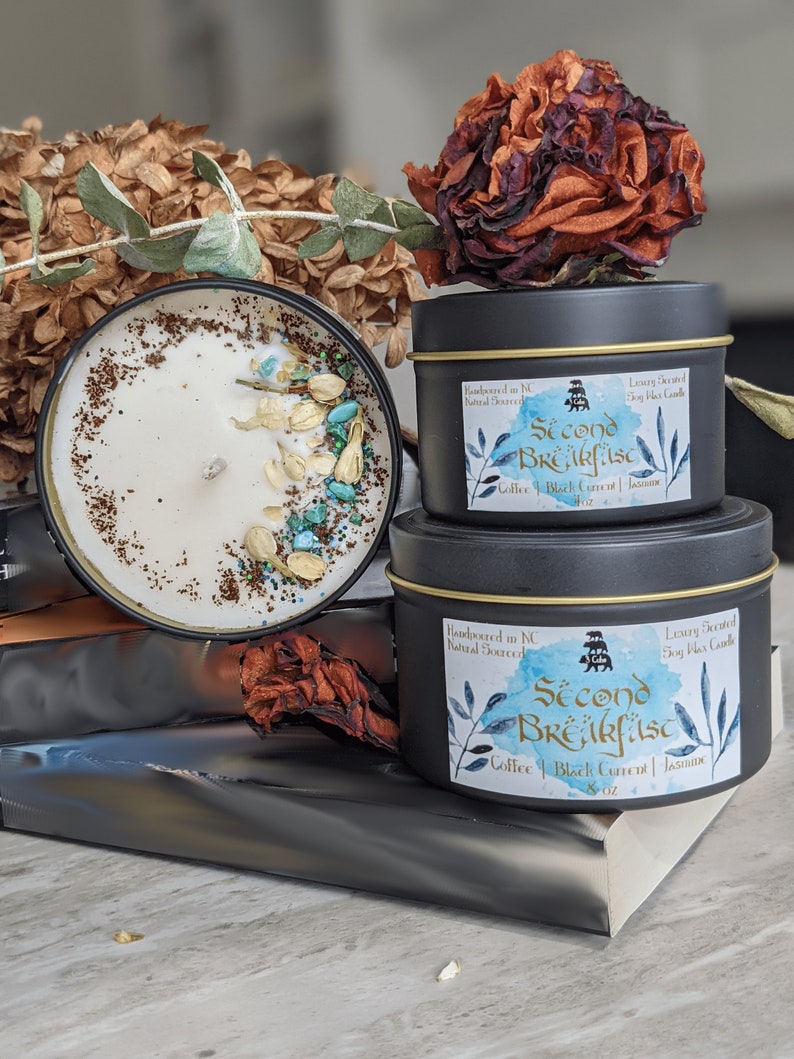 May include: Two black candle jars with gold lids. The smaller jar is on top of the larger jar. Both jars have a white label with black text that reads "Second Breakfast" and "Coffee | Black Current | Jasmine". The larger jar is 8 oz. The smaller jar is 4 oz.