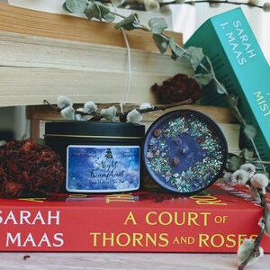 May include: A black candle tin with a blue and silver label that reads "Night Triumphant" with a star-filled background. The candle is sitting on a red book with the title "A Court of Thorns and Roses" by Sarah J. Maas.