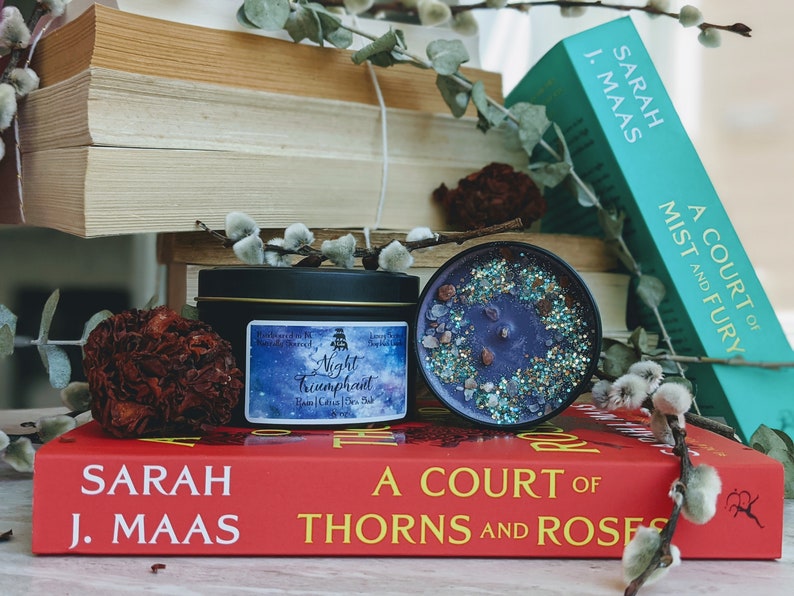 May include: A black candle tin with a blue and silver label that reads "Night Triumphant" with a star-filled background. The candle is sitting on a red book with the title "A Court of Thorns and Roses" by Sarah J. Maas.