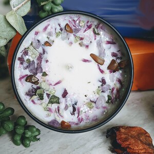 May include: A white candle with a purple and green floral design and embedded crystals. The candle is in a black container.