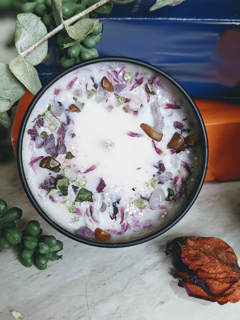 May include: A white candle with a purple and green floral design and embedded crystals. The candle is in a black container.