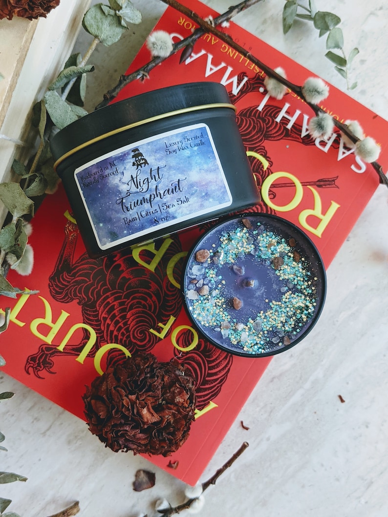 A beautifully crafted candle labeled "Night Triumphant," featuring a luxurious blend of rain, citrus, and sea salt scents. The candle is placed on a vibrant red book titled "A Court of Mist and Fury" by Sarah J. Maas, surrounded by dried flowers and decorative elements. The candle's surface is adorned with sparkling blue and silver glitter, resembling a starry night, adding a magical touch to the display.