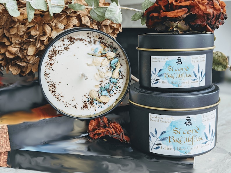 A beautifully crafted candle named "Second Breakfast" displayed among dried flowers. The candle features a creamy white wax topped with ground coffee, floral jasmine petals, and turquoise crystal shards. Two black metal tins with gold accents sit beside it, each labeled with the candle's name and scent notes of coffee, black currant, and jasmine.