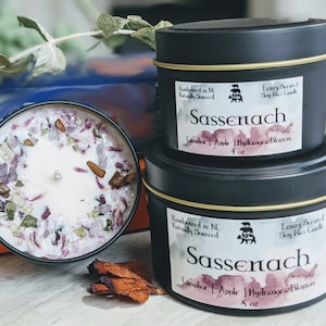 Two Sassenach candles are displayed, one in a 4oz black metal tin and the other in an 8oz jar. Both feature a beautiful floral and crystal embellishment on top, showcasing vibrant hues of purple and earthy tones. The labels highlight the scent notes of Camphor, Apple, and Hydrangea Blossom.