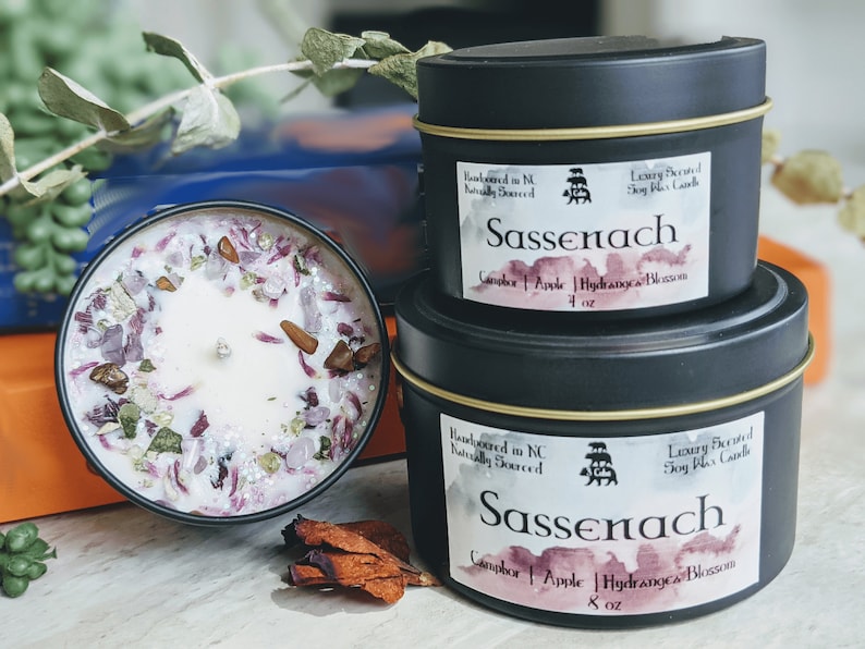 Two Sassenach candles are displayed, one in a 4oz black metal tin and the other in an 8oz jar. Both feature a beautiful floral and crystal embellishment on top, showcasing vibrant hues of purple and earthy tones. The labels highlight the scent notes of Camphor, Apple, and Hydrangea Blossom.