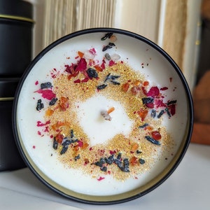 K&ouml;nnte beinhalten: Eine wei&szlig;e Kerze mit schwarzem Rand, bedeckt mit einer dekorativen Schicht aus goldenem Glitzer, getrockneten Rosenbl&auml;ttern und kleinen schwarzen und orangefarbenen St&uuml;cken.