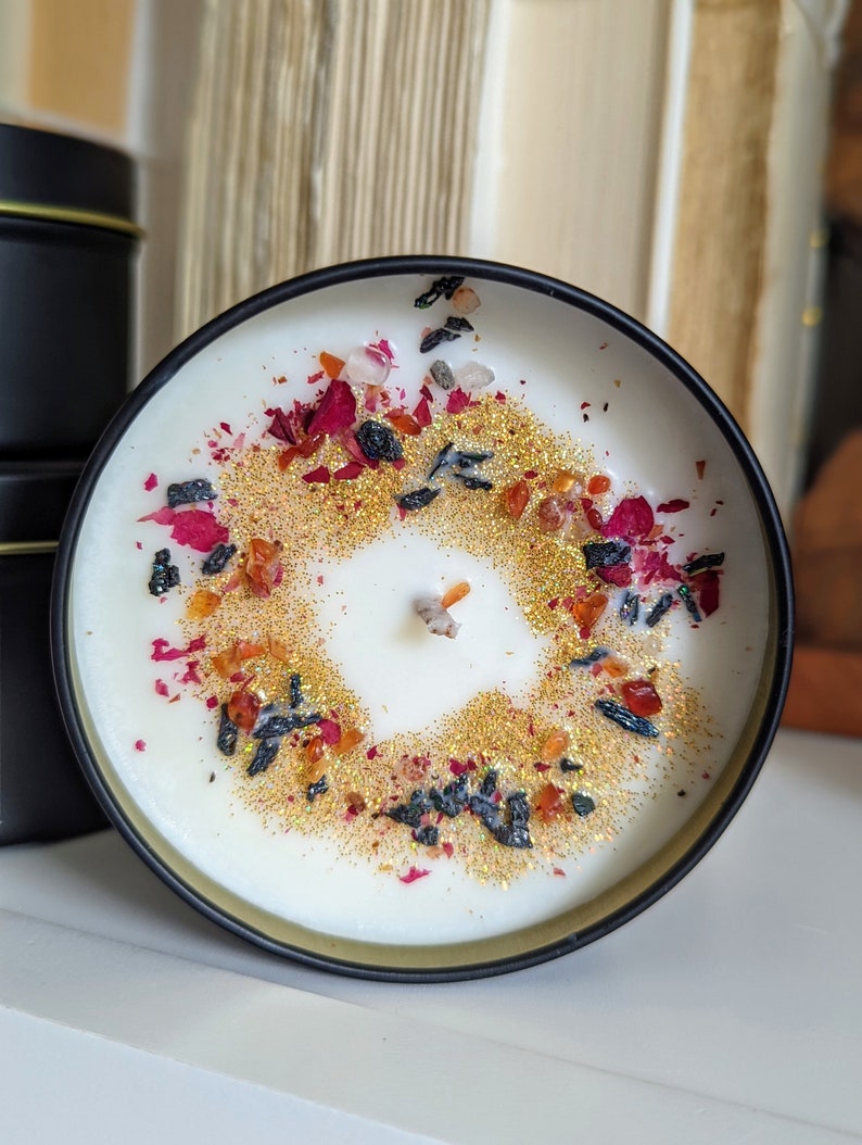 K&ouml;nnte beinhalten: Eine wei&szlig;e Kerze mit schwarzem Rand, bedeckt mit einer dekorativen Schicht aus goldenem Glitzer, getrockneten Rosenbl&auml;ttern und kleinen schwarzen und orangefarbenen St&uuml;cken.