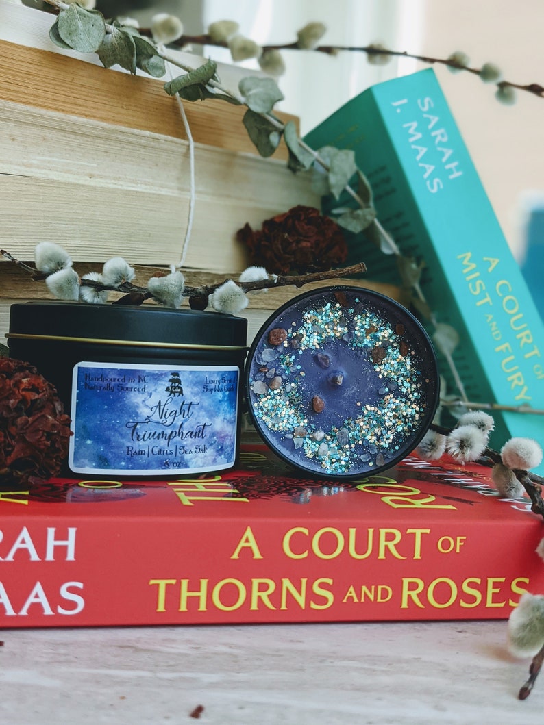 May include: A black candle jar with a purple wax and glitter on top. The candle jar has a label that reads "Night Triumphant" with the scent notes "Ram, Citrus, Sea Salt". The candle jar is sitting on top of a red book with the title "A Court of Thorns and Roses".