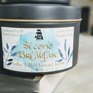 May include: A black candle tin with a gold lid and a white label. The label has a watercolor design with blue and green leaves. The text on the label reads "Second Breakfast" in gold lettering, "Coffee | Black Current | Jasmine" in black lettering, and "8 oz" in black lettering. The label also includes a logo with three bears stacked on top of each other.