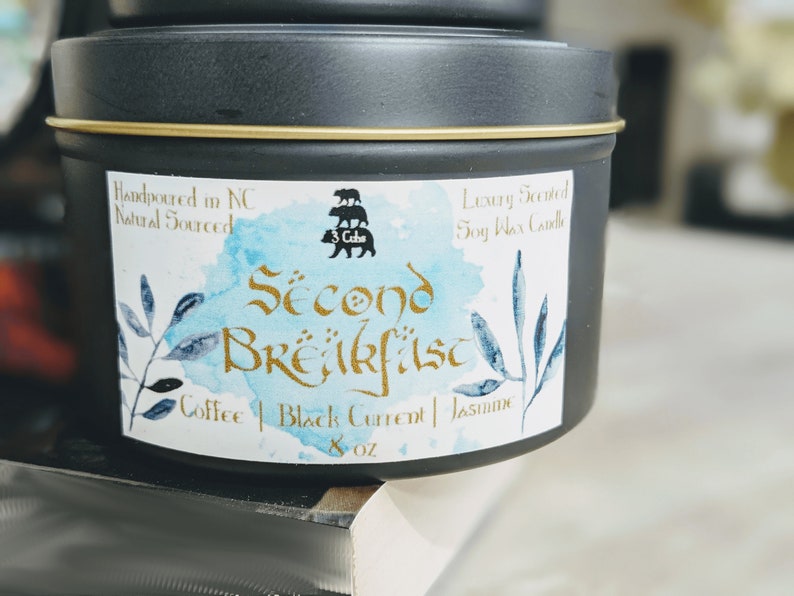 May include: A black candle tin with a gold lid and a white label. The label has a watercolor design with blue and green leaves. The text on the label reads "Second Breakfast" in gold lettering, "Coffee | Black Current | Jasmine" in black lettering, and "8 oz" in black lettering. The label also includes a logo with three bears stacked on top of each other.