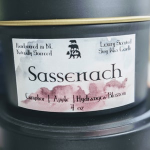 May include: A black tin candle with a white label that says "Handpoured in NC Naturally Sourced" and "Luxury Scented Soy Wax Candle". The candle is labeled "Sassenach" and has a scent description of "Camphor | Apple | Hydrangea Blossom" and is 4 oz.