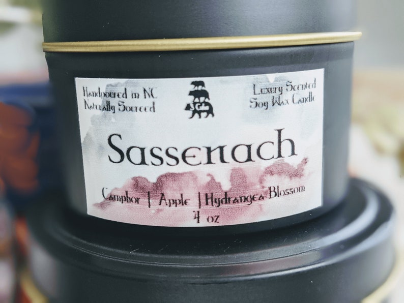 May include: A black tin candle with a white label that says "Handpoured in NC Naturally Sourced" and "Luxury Scented Soy Wax Candle". The candle is labeled "Sassenach" and has a scent description of "Camphor | Apple | Hydrangea Blossom" and is 4 oz.