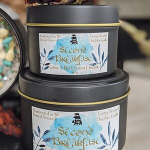 May include: Two black metal candle tins with gold lids. The labels are blue and white with a watercolor effect. The larger tin is labeled "Second Breakfast" with the text "Coffee | Black Current | Jasmine" and "8 oz". The smaller tin is labeled "Second Breakfast" with the text "Coffee | Black Current | Jasmine" and "4 oz". Both tins are hand-poured in North Carolina with natural soy wax.