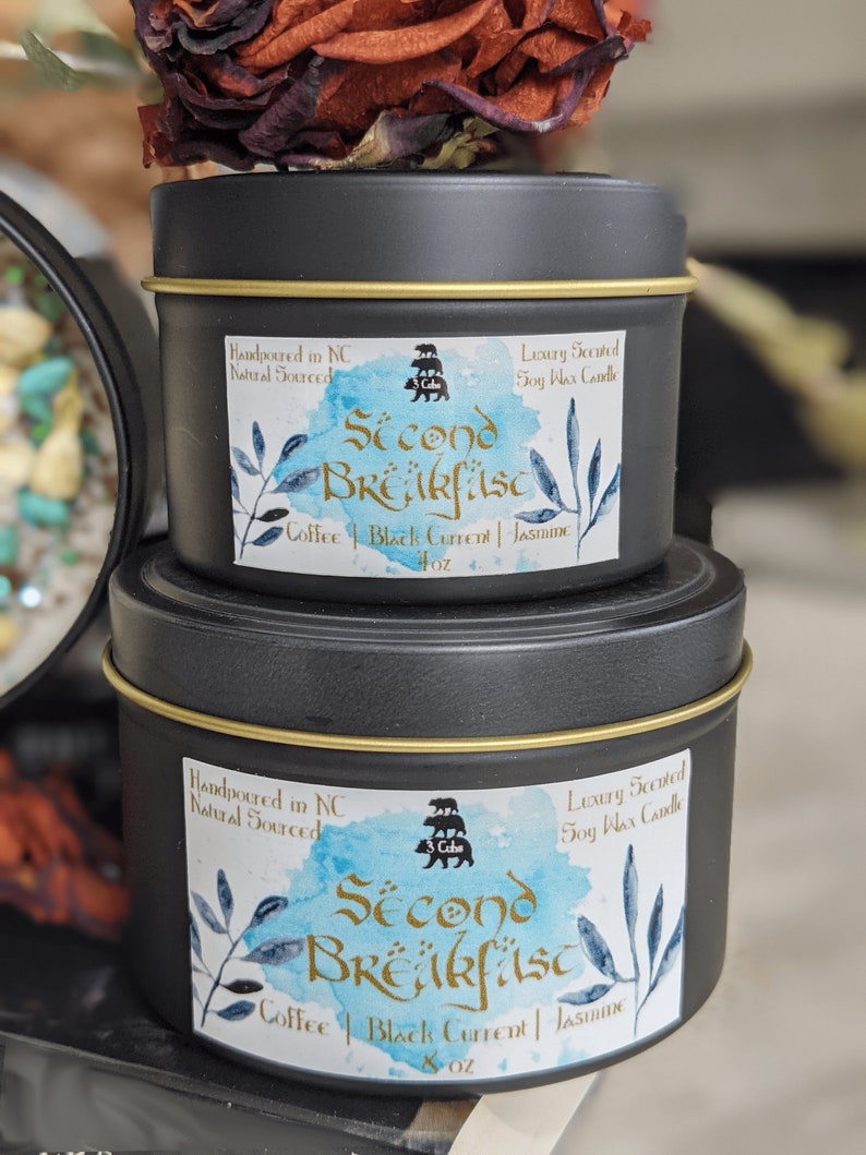 May include: Two black metal candle tins with gold lids. The labels are blue and white with a watercolor effect. The larger tin is labeled "Second Breakfast" with the text "Coffee | Black Current | Jasmine" and "8 oz". The smaller tin is labeled "Second Breakfast" with the text "Coffee | Black Current | Jasmine" and "4 oz". Both tins are hand-poured in North Carolina with natural soy wax.