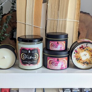K&ouml;nnte beinhalten: Drei Kerzen in verschiedenen Gr&ouml;&szlig;en mit schwarzen Deckeln und Etiketten, auf denen "Starborn" in rosa und wei&szlig;en Buchstaben steht. Die Kerzen stehen auf einem wei&szlig;en Regal mit B&uuml;chern im Hintergrund.