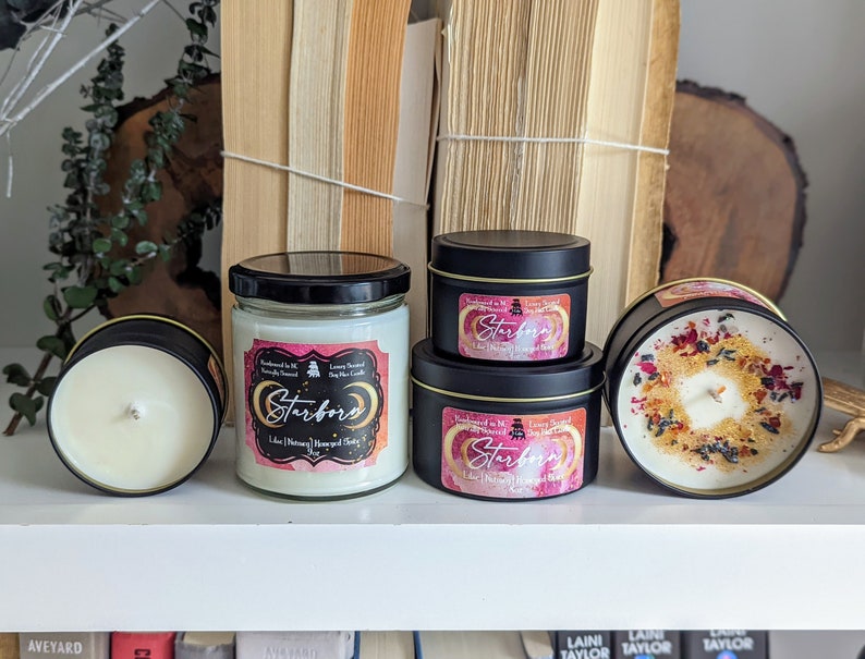 K&ouml;nnte beinhalten: Drei Kerzen in verschiedenen Gr&ouml;&szlig;en mit schwarzen Deckeln und Etiketten, auf denen "Starborn" in rosa und wei&szlig;en Buchstaben steht. Die Kerzen stehen auf einem wei&szlig;en Regal mit B&uuml;chern im Hintergrund.