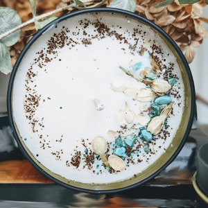 May include: A white candle with a brown and blue speckled rim. The candle is in a black tin container with a gold rim. The candle is decorated with white flower petals and blue and green stones.