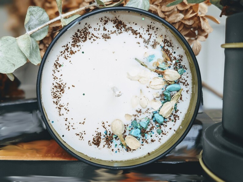 May include: A white candle with a brown and blue speckled rim. The candle is in a black tin container with a gold rim. The candle is decorated with white flower petals and blue and green stones.