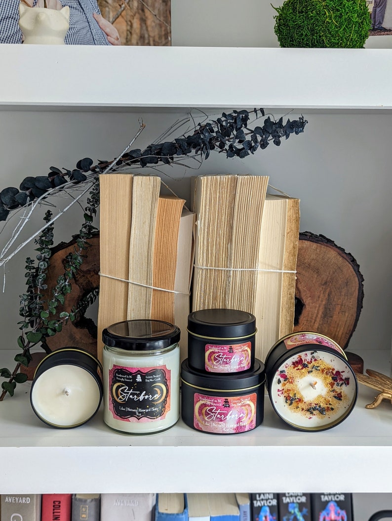K&ouml;nnte beinhalten: Drei schwarze Kerzenbeh&auml;lter mit Deckeln auf einem wei&szlig;en Regal. Die Kerzen sind mit "Starborn" beschriftet und haben ein rosafarbenes und goldenes Design. Die Kerzen sind von B&uuml;chern und Gr&uuml;npflanzen umgeben.