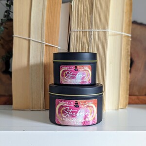 K&ouml;nnte beinhalten: Zwei schwarze Metall-Kerzenbeh&auml;lter mit goldenen Deckeln. Die Etiketten sind rosa und goldfarben mit dem Text "Starborn Luxury Scented Soy Blend Candle" und einer Grafik eines Halbmondes.