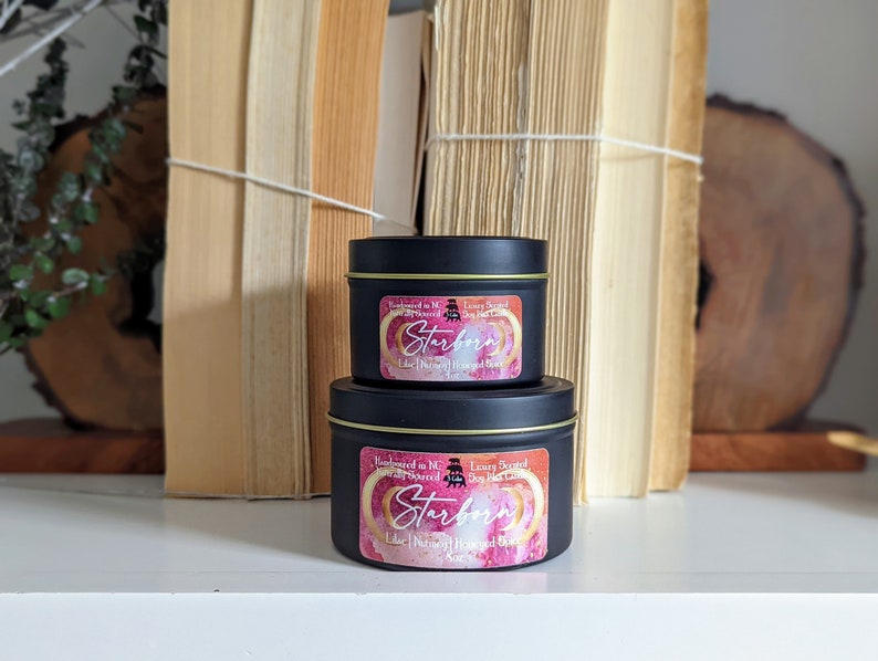 K&ouml;nnte beinhalten: Zwei schwarze Metall-Kerzenbeh&auml;lter mit goldenen Deckeln. Die Etiketten sind rosa und goldfarben mit dem Text "Starborn Luxury Scented Soy Blend Candle" und einer Grafik eines Halbmondes.