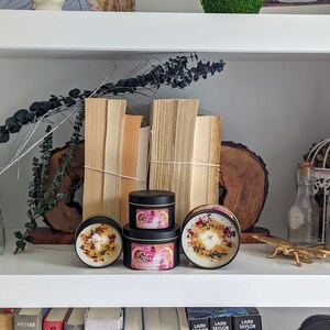 K&ouml;nnte beinhalten: Drei schwarze Kerzenbeh&auml;lter mit Blumendesigns und dem Text "Starfire" auf der Vorderseite. Die Kerzen stehen auf einem wei&szlig;en Regal mit B&uuml;chern, Pflanzen und Dekorationsartikeln.