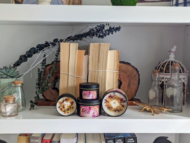 K&ouml;nnte beinhalten: Drei schwarze Kerzenbeh&auml;lter mit Blumendesigns und dem Text "Starfire" auf der Vorderseite. Die Kerzen stehen auf einem wei&szlig;en Regal mit B&uuml;chern, Pflanzen und Dekorationsartikeln.