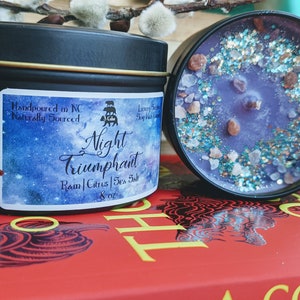 May include: A black candle tin with a white label that reads "Night Triumphant" with the scent notes "Rain | Citrus | Sea Salt" and a 8 oz size. The candle is hand-poured in North Carolina and naturally sourced. The candle is sitting on a red book with a black design.