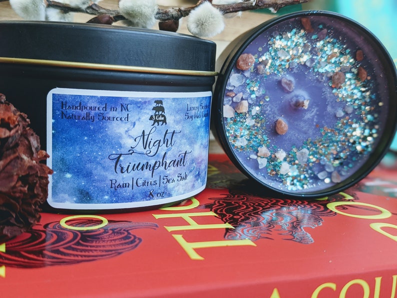 May include: A black candle tin with a white label that reads "Night Triumphant" with the scent notes "Rain | Citrus | Sea Salt" and a 8 oz size. The candle is hand-poured in North Carolina and naturally sourced. The candle is sitting on a red book with a black design.