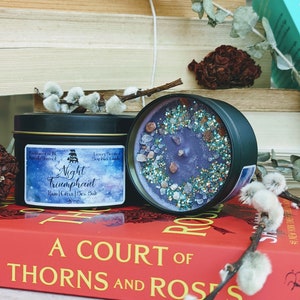 May include: A purple soy wax candle with a glittery, blue and gold finish. The candle is in a black tin with the text "Night Triumphant" and "Rain | Citrus | Sea Salt" on the label. The candle is sitting on a book titled "A Court of Thorns and Roses".
