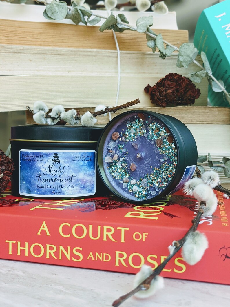 May include: A purple soy wax candle with a glittery, blue and gold finish. The candle is in a black tin with the text "Night Triumphant" and "Rain | Citrus | Sea Salt" on the label. The candle is sitting on a book titled "A Court of Thorns and Roses".