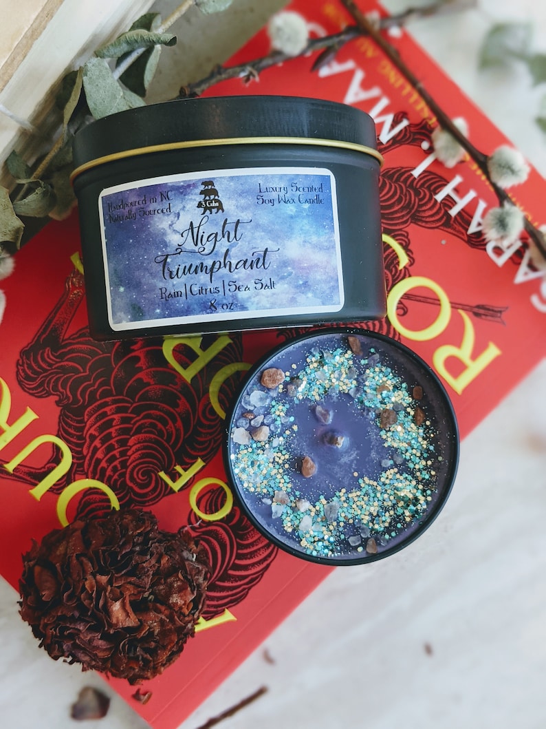 May include: A black soy wax candle with a gold lid. The label reads "Handpoured in NC Naturally Sourced" and "Luxury Scented Soy Wax Candle". The candle is labeled "Night Triumphant" with the scent notes "Rain | Citrus | Sea Salt" and is 8 oz.