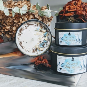 May include: Two black candle jars with gold lids. The smaller jar is on top of the larger jar. Both jars have a blue and white label with the text "Second Breakfast" and the words "Coffee, Black Currant & Jasmine".