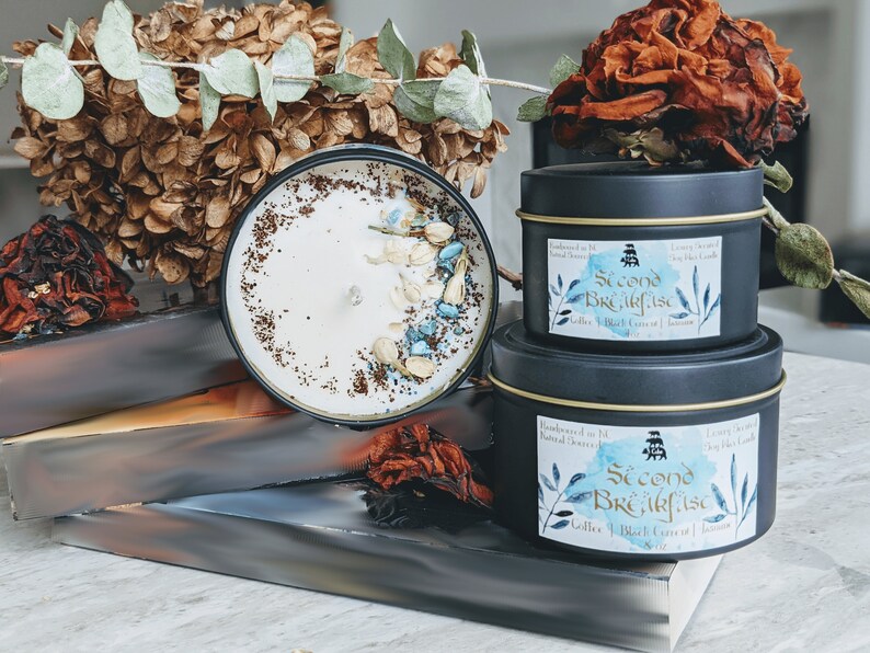 May include: Two black candle jars with gold lids. The smaller jar is on top of the larger jar. Both jars have a blue and white label with the text "Second Breakfast" and the words "Coffee, Black Currant & Jasmine".