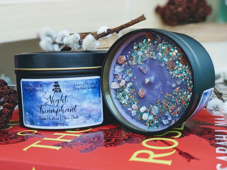 May include: A purple soy wax candle in a black tin with a galaxy design. The candle is labeled "Night Triumphant" and has a scent of Rain, Citrus, and Sea Salt. The candle is 8 oz.