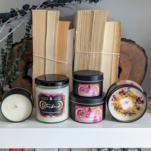 K&ouml;nnte beinhalten: Drei schwarze und goldene Kerzen mit dem Etikett "Starborn" und einem rosafarbenen und wei&szlig;en Aquarelldesign. Die Kerzen stehen auf einem wei&szlig;en Regal mit B&uuml;chern im Hintergrund.