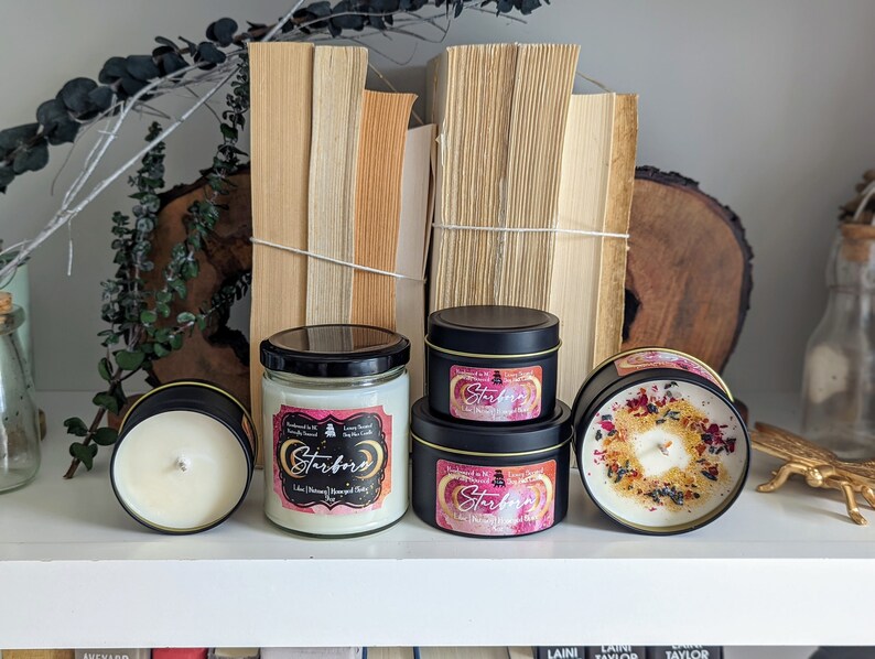 K&ouml;nnte beinhalten: Drei schwarze und goldene Kerzen mit dem Etikett "Starborn" und einem rosafarbenen und wei&szlig;en Aquarelldesign. Die Kerzen stehen auf einem wei&szlig;en Regal mit B&uuml;chern im Hintergrund.