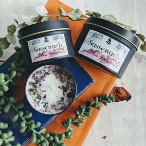 May include: Two black candle tins with gold lids and labels that read "Sassenach" in a white script. The candles are labeled "Lavender & Pink" and "Hydrangea & Blossom" and are 4 oz each. One candle is lit and the wax is melted, revealing pink and green flower petals.
