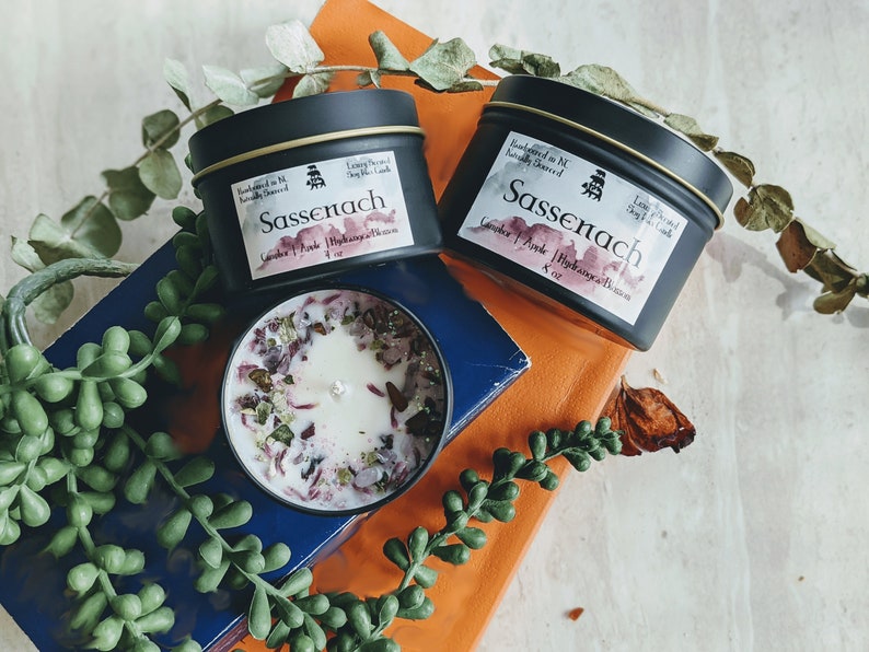 May include: Two black candle tins with gold lids and labels that read "Sassenach" in a white script. The candles are labeled "Lavender & Pink" and "Hydrangea & Blossom" and are 4 oz each. One candle is lit and the wax is melted, revealing pink and green flower petals.