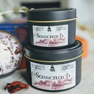 May include: Two black candle tins with gold lids. The larger tin is labeled "Sassenach" with the text "Handpoured in NC Naturally Sourced" and "Luxury Scented Soy Wax Candle". The smaller tin is labeled "Sassenach" with the text "Handpoured in NC Naturally Sourced" and "Luxury Scented Soy Wax Candle". Both tins have the text "Camphor | Apple | Hydrangea Blossom" and the size in ounces.