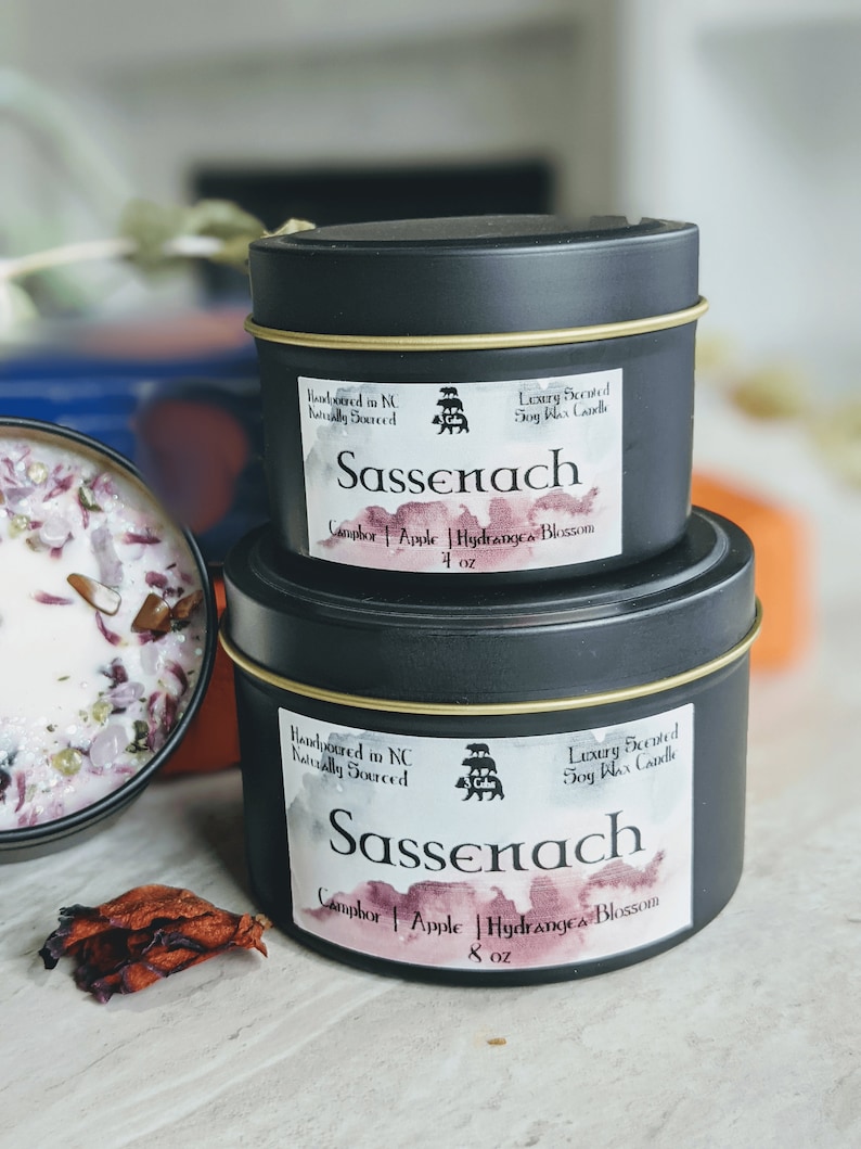May include: Two black candle tins with gold lids. The larger tin is labeled "Sassenach" with the text "Handpoured in NC Naturally Sourced" and "Luxury Scented Soy Wax Candle". The smaller tin is labeled "Sassenach" with the text "Handpoured in NC Naturally Sourced" and "Luxury Scented Soy Wax Candle". Both tins have the text "Camphor | Apple | Hydrangea Blossom" and the size in ounces.