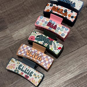 May include: Six colorful hair clips with different patterns and names printed on them. The names are "Sophia", "Bella", "Mia", "Emma", "Olivia", and "Ellie".