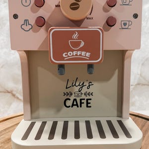 May include: A pink wooden toy coffee maker with a brown accent panel that reads "COFFEE". The coffee maker has several pink buttons, a dial with coffee bean icons, and the text "Lily's CAFE". The base is white with brown stripes.